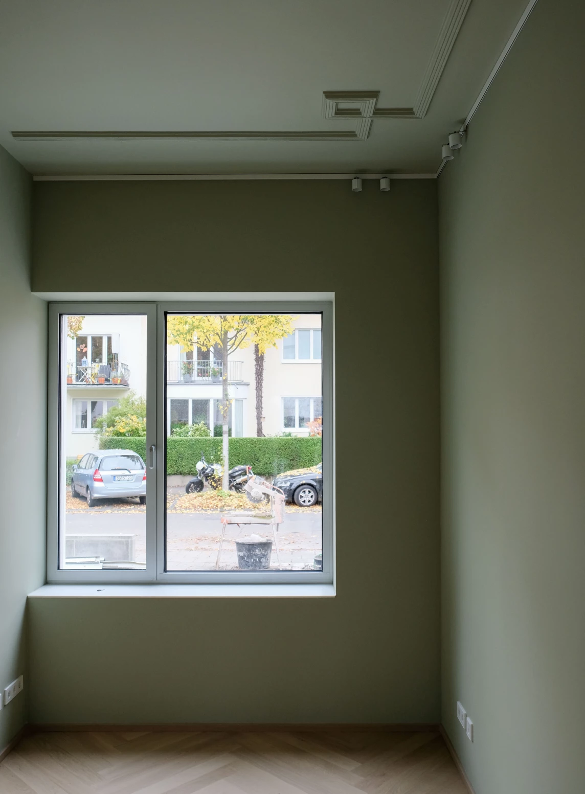 Ein heller Raum mit einem Fenster, das auf eine herbstliche Straße mit Autos und einem Baum hinausblickt.