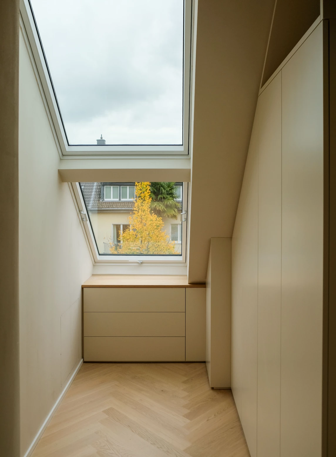 Ein heller Raum mit Dachfenster und Blick auf einen herbstlichen Baum vor Wohngebäuden.