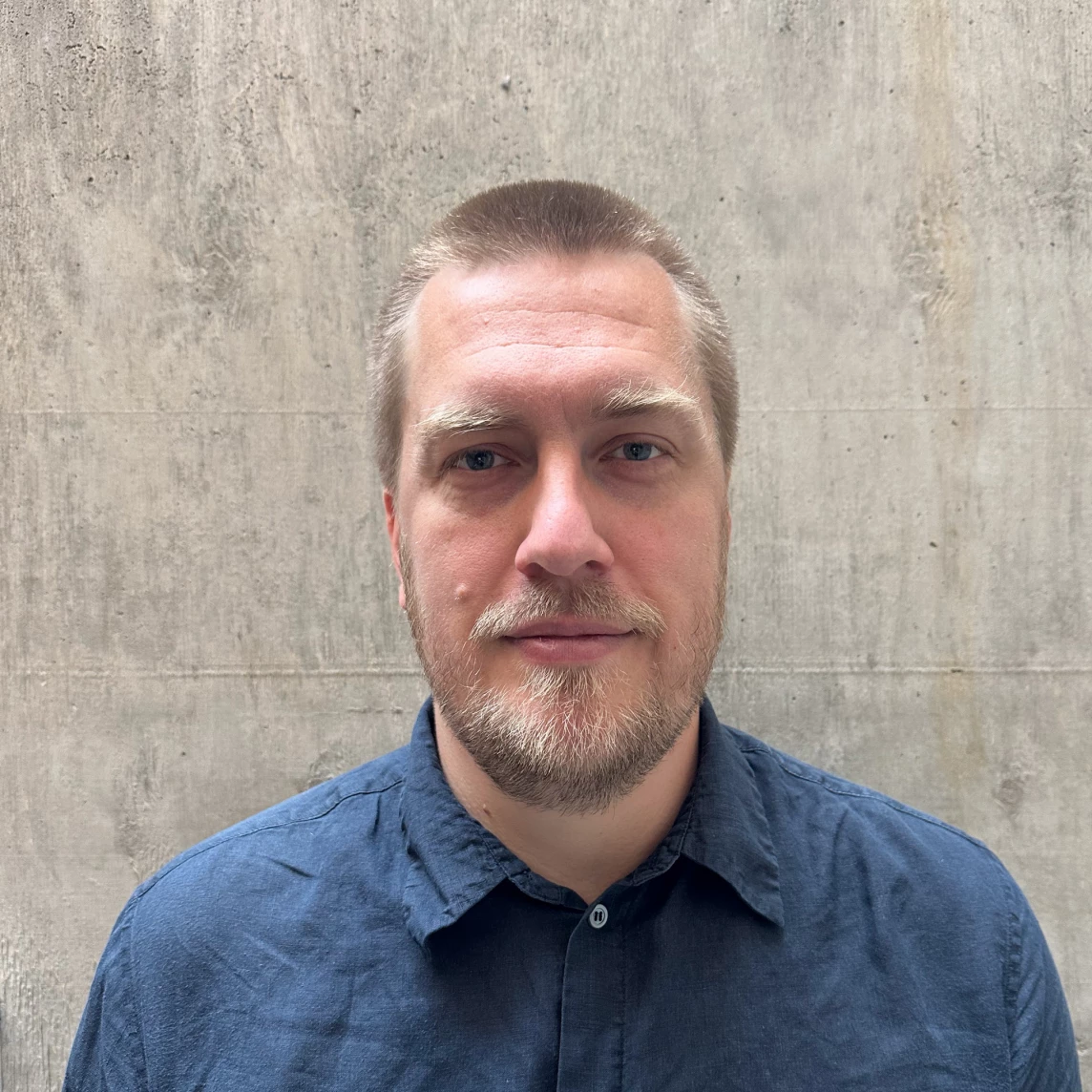 Ein Mann mit kurzem, blondem Haar und einem Bart steht vor einer grauen Wand.