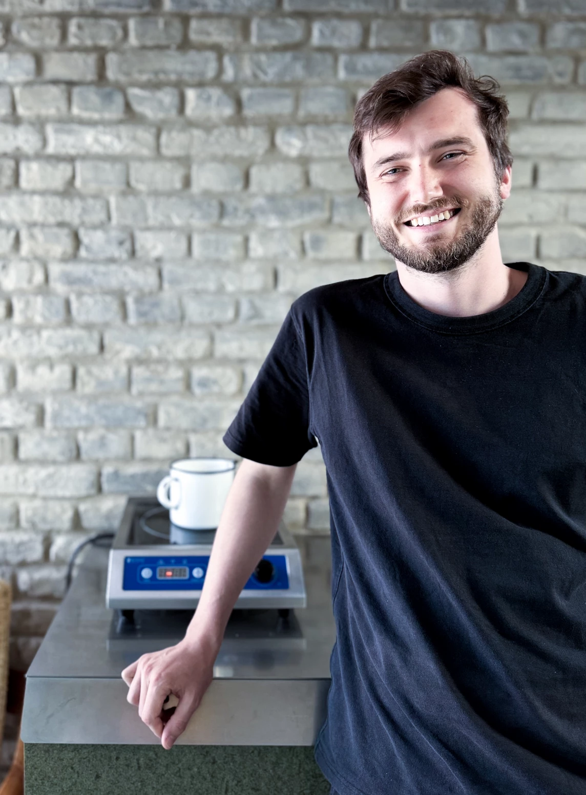 Ein lächelnder junger Mann steht am Tisch mit einer Kochplatte und einer Tasse auf einem Marmoruntergrund.