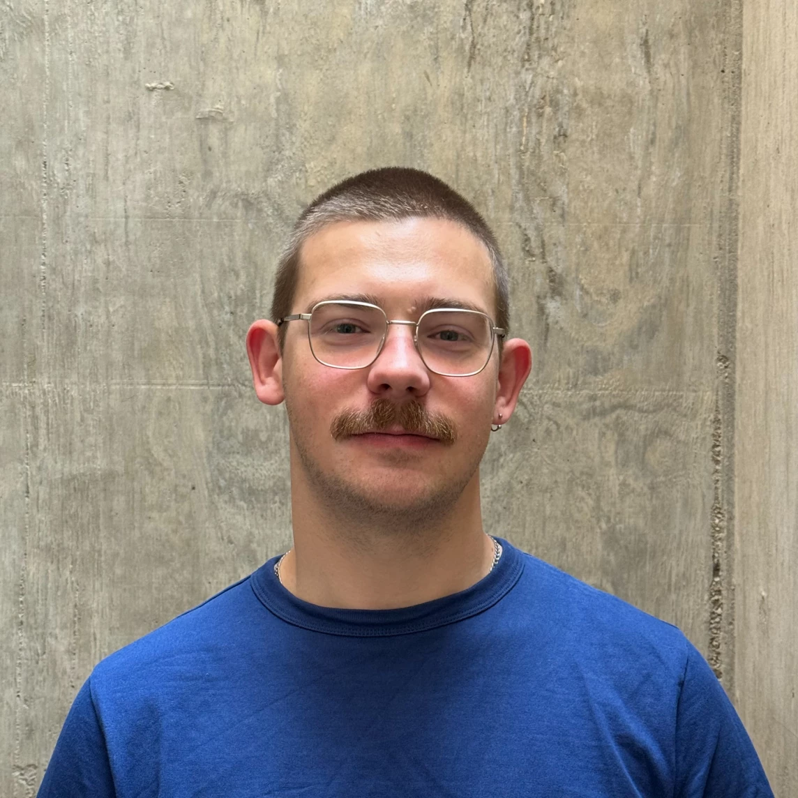 Ein junger Mann mit Brille und Schnurrbart, trägt ein blaues T-Shirt vor einer grauen Wand.