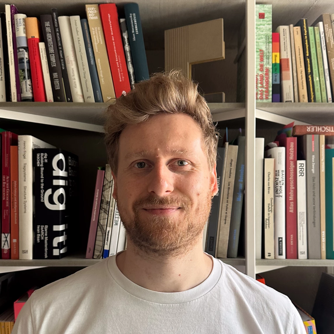 Ein Mensch mit blonden Haaren lächelt vor einem Bücherregal mit vielen farbigen Büchern.