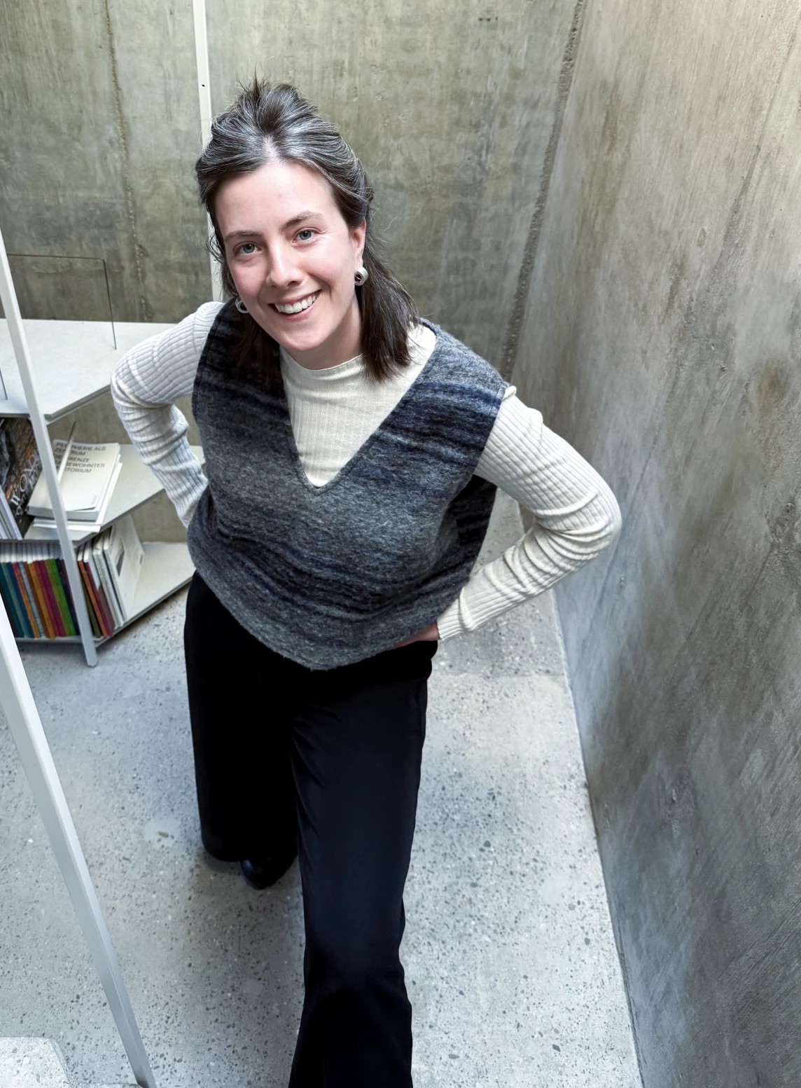Eine Person mit dunklem Haar, die in einem modernen Raum in einem grauen Oberteil und schwarzer Hose posiert.