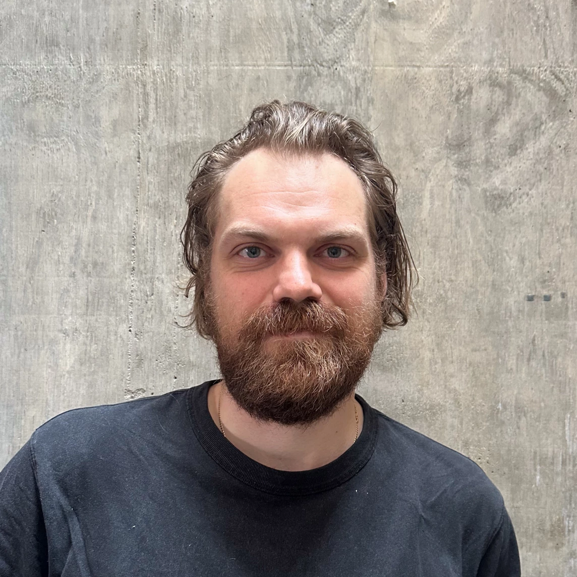 Ein Mann mit Bart und lockigem Haar, trägt ein schwarzes T-Shirt, vor einer grauen Wand.