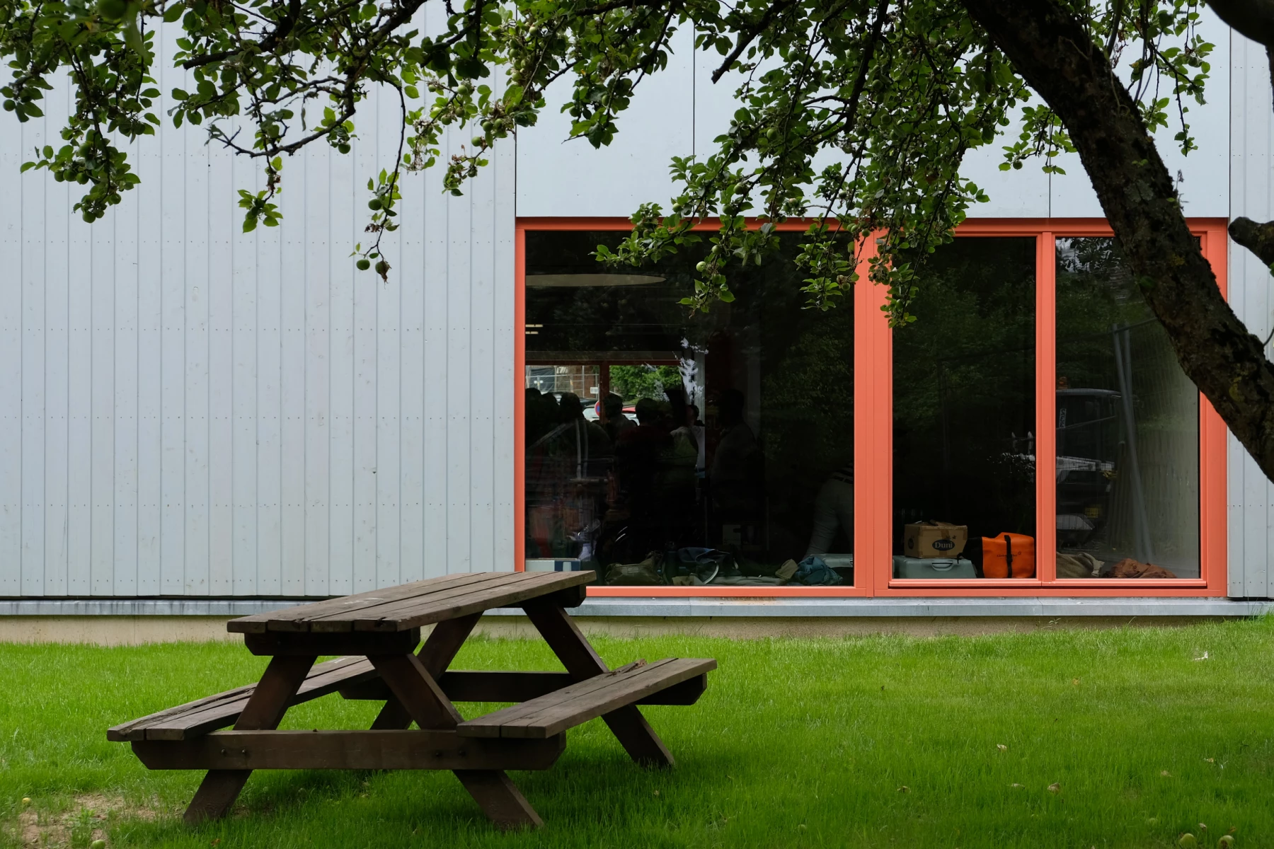 Ein Holztisch auf einer grünen Wiese vor einem modernen, orange eingerahmten Fenster.