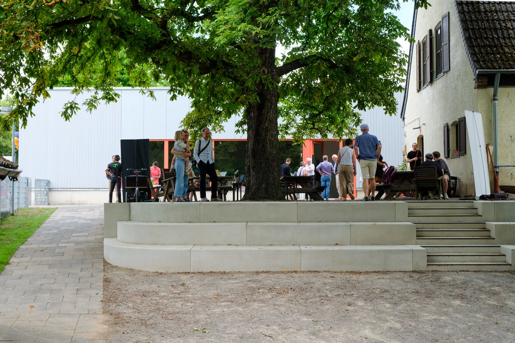 Eine Gruppe von Menschen versammelt sich unter einem Baum vor einem Gebäude mit orangefarbenen Fenstern.