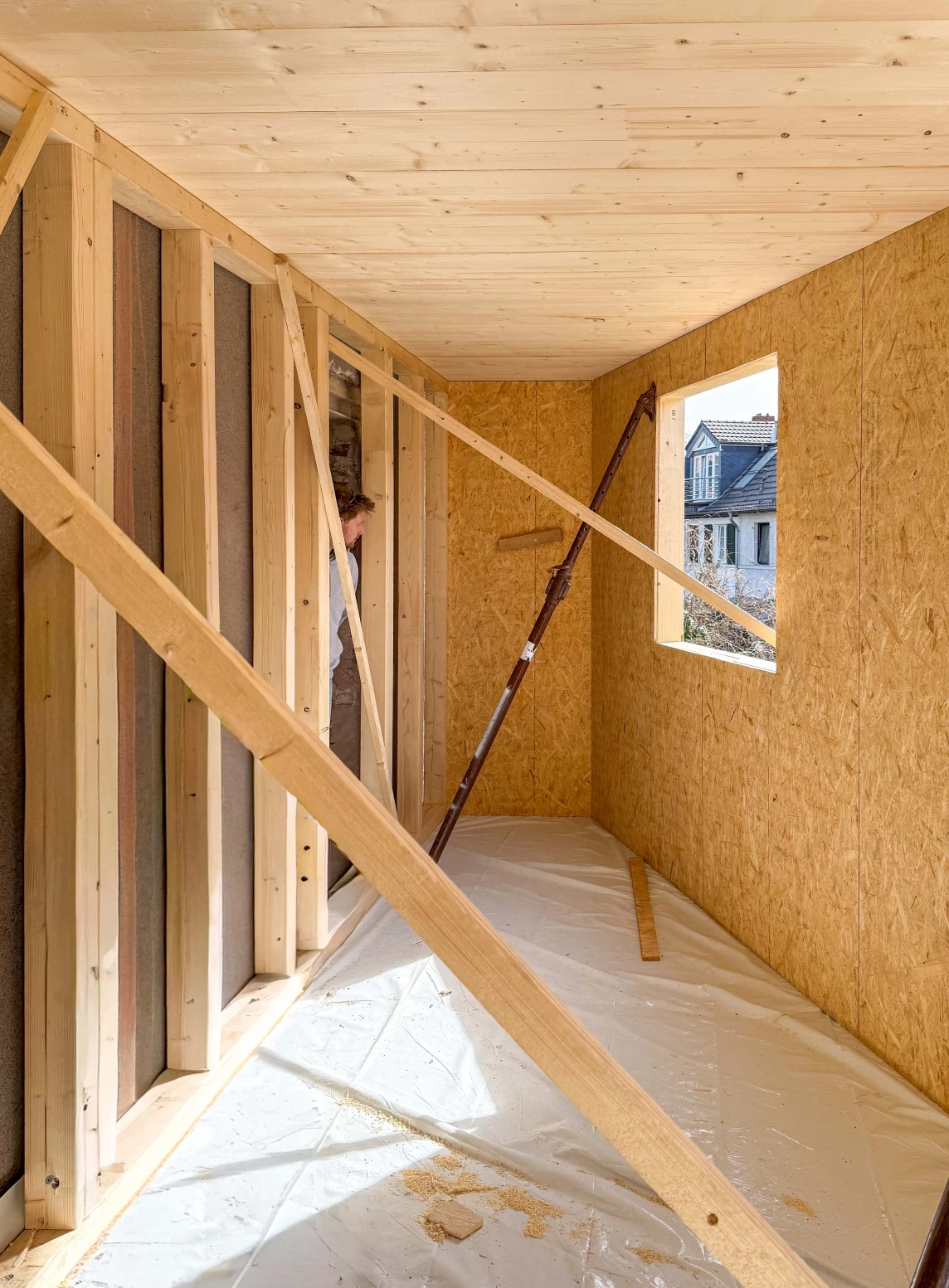Innenansicht eines Rohbaus mit Holzrahmen, Fenster und einem Arbeiter, der mit Bauarbeiten beschäftigt ist.