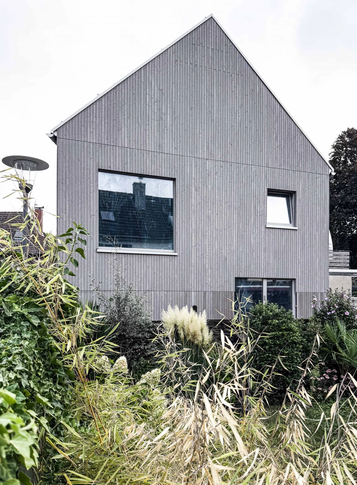Ein modernes, graues Holzhaus mit großen Fenstern, umgeben von üppigem, grünem Garten und dekorativen Pflanzen.
