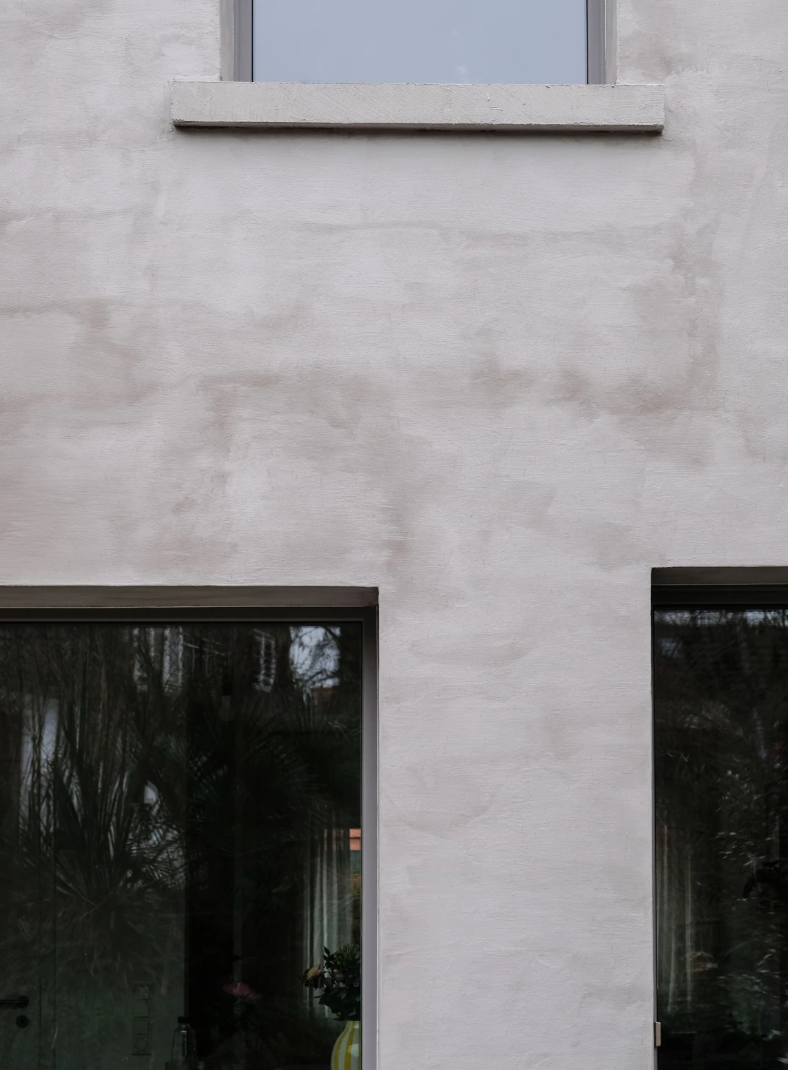 Die Wand eines Hauses mit zwei großen Fenstern und einem dezenten Putz in hellen Farbtönen.